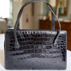 May include: A black, rectangular handbag with a crocodile skin texture. The bag has two curved handles and a zipper closure. The bag is sitting on a white surface.