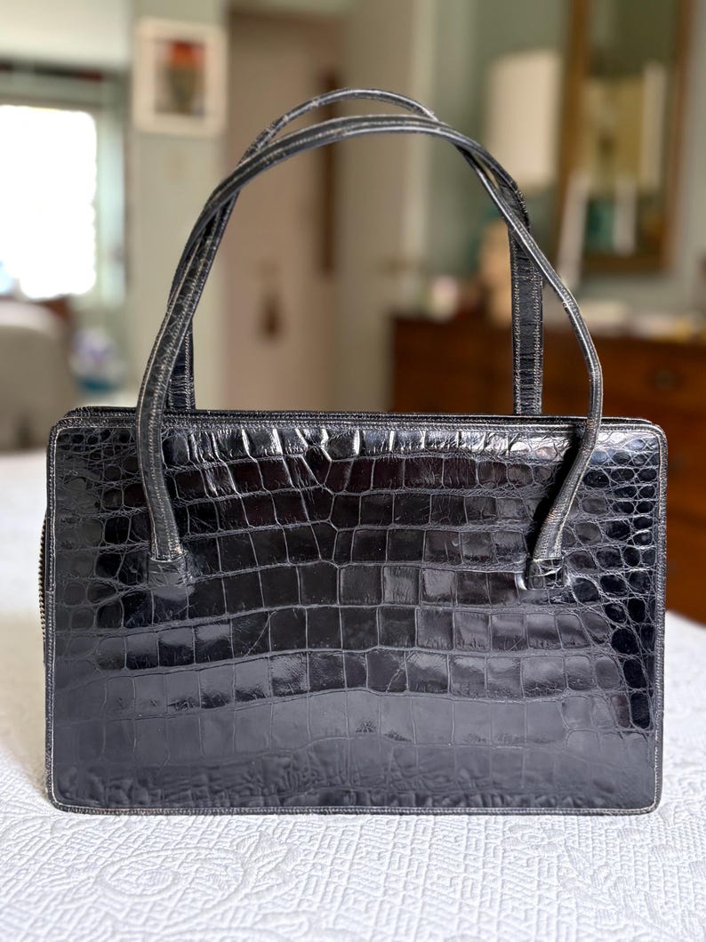 May include: A black, rectangular handbag with a crocodile skin texture. The bag has two curved handles and a zipper closure. The bag is sitting on a white surface.