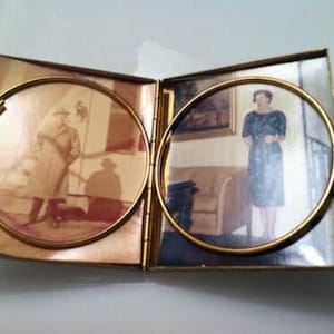 May include: A gold-toned hinged photo frame with two round openings. The frame contains two black and white photos. One photo shows a person wearing a coat and hat, and the other photo shows a person wearing a patterned dress.