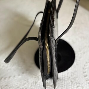 May include: Black handbag with a textured exterior and a gold-colored zipper. The bag has thin, black straps and a rectangular shape. The bag is displayed on a black stand against a white patterned background.