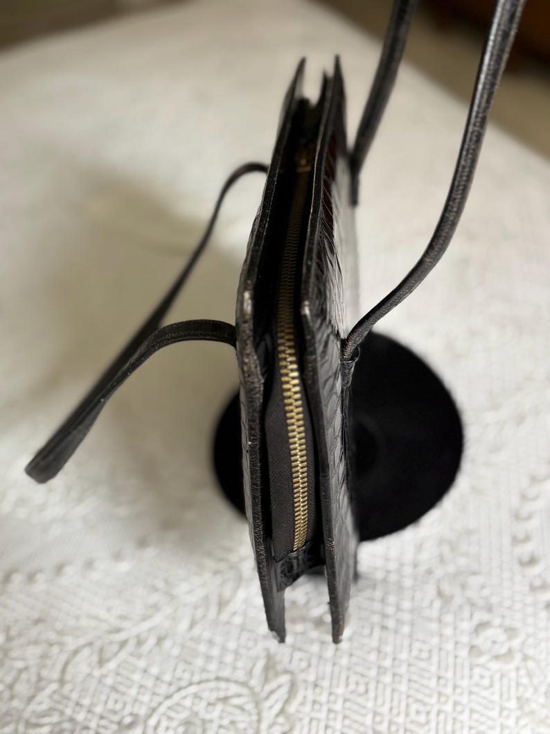 May include: Black handbag with a textured exterior and a gold-colored zipper. The bag has thin, black straps and a rectangular shape. The bag is displayed on a black stand against a white patterned background.