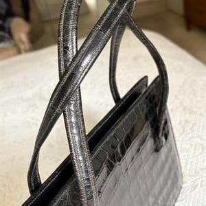 May include: A black handbag with a crocodile-like texture. The bag has two handles and a rectangular shape. The handles are thin and curved, and the bag appears to be made of leather or a similar material. The bag is sitting on a white surface.