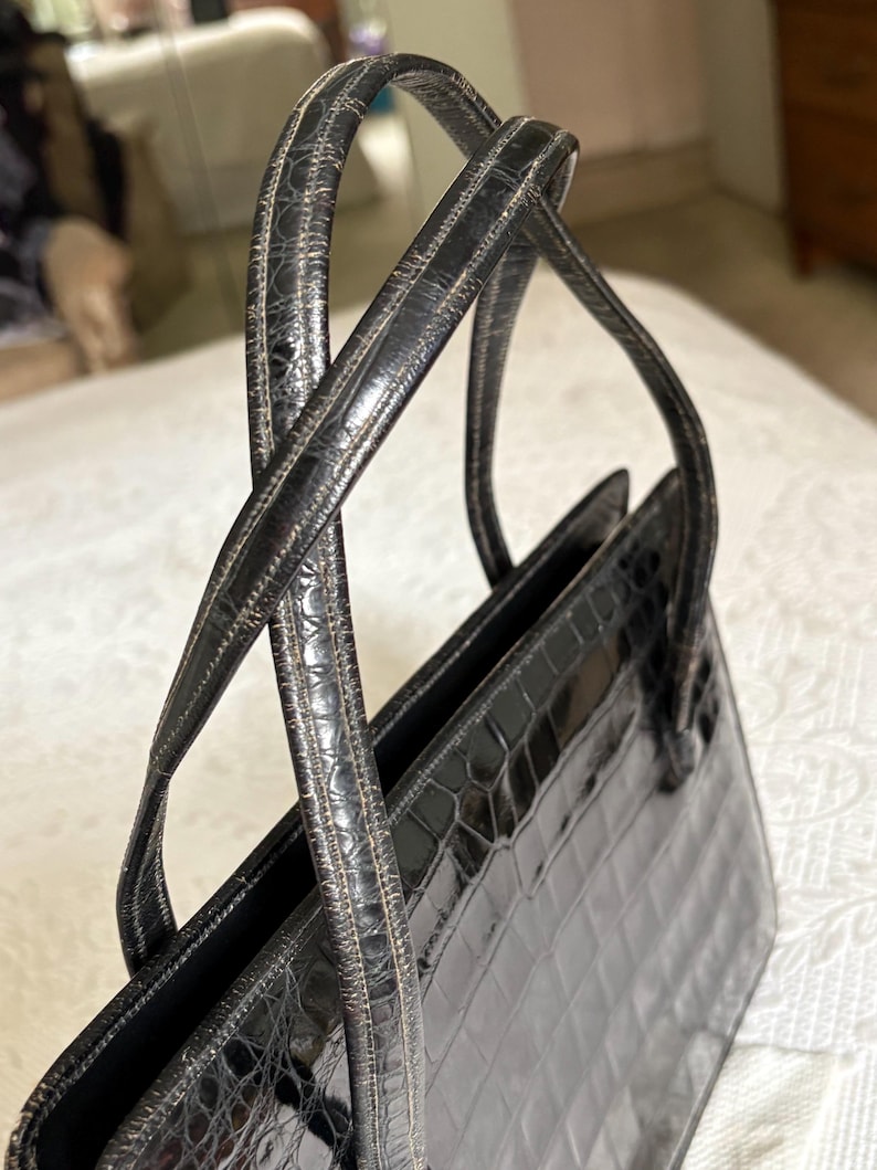 May include: A black handbag with a crocodile-like texture. The bag has two handles and a rectangular shape. The handles are thin and curved, and the bag appears to be made of leather or a similar material. The bag is sitting on a white surface.