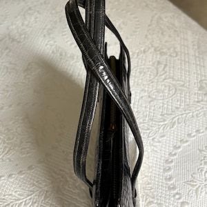 May include: Black leather tote bag with a crocodile skin texture. The bag has two handles and a zipper closure. The bag is made of a shiny, dark material.