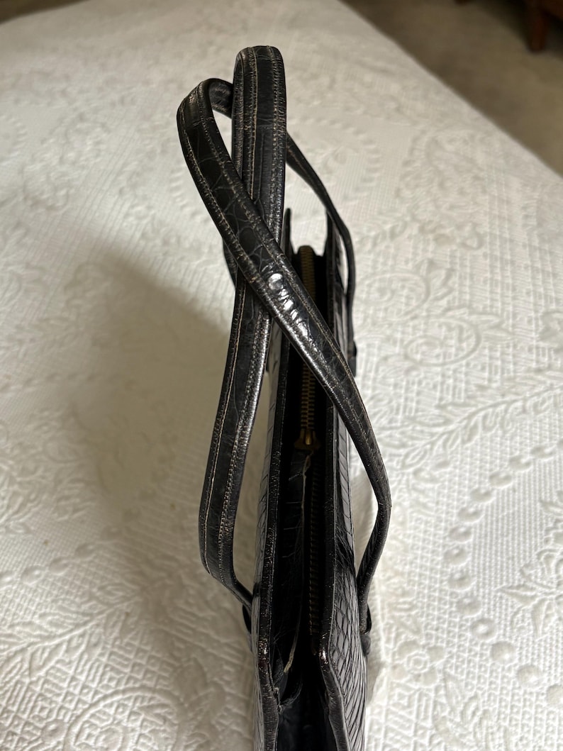 May include: Black leather tote bag with a crocodile skin texture. The bag has two handles and a zipper closure. The bag is made of a shiny, dark material.