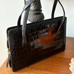 May include: A black, rectangular handbag with a crocodile skin texture. The bag has two handles and a gold-colored zipper. The handbag is sitting on a wooden surface.