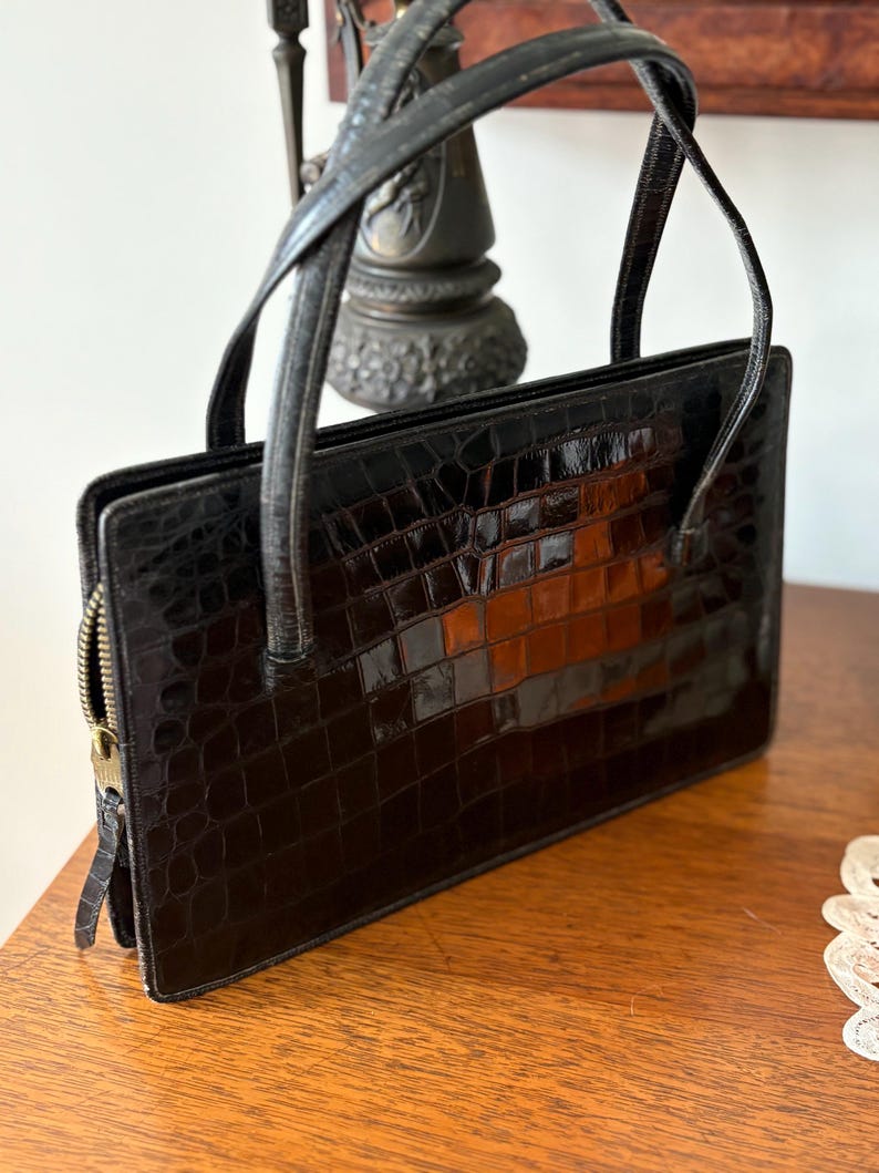 May include: A black, rectangular handbag with a crocodile skin texture. The bag has two handles and a gold-colored zipper. The handbag is sitting on a wooden surface.