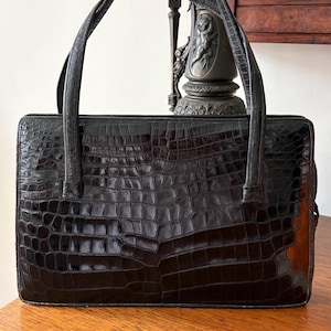 May include: Black rectangular handbag with a crocodile skin pattern. The bag has two curved handles and a rectangular shape. The bag is sitting on a wooden surface. The bag is a dark, glossy black color.