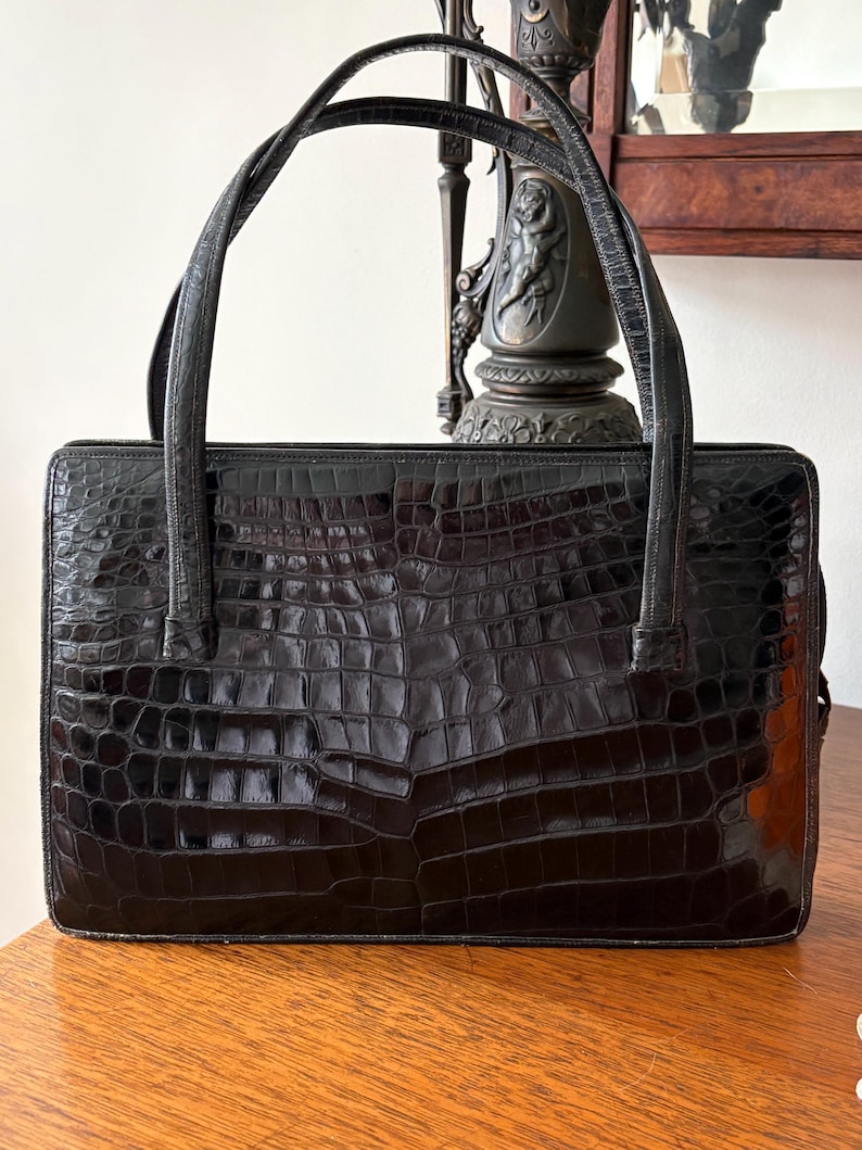 May include: Black rectangular handbag with a crocodile skin pattern. The bag has two curved handles and a rectangular shape. The bag is sitting on a wooden surface. The bag is a dark, glossy black color.
