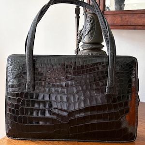 May include: A black, rectangular handbag with a crocodile skin texture. The bag has two curved handles and a structured design. The glossy finish reflects light, highlighting the embossed pattern. The bag sits on a wooden surface.
