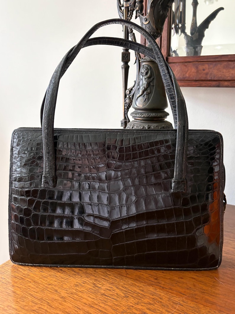 May include: A black, rectangular handbag with a crocodile skin texture. The bag has two curved handles and a structured design. The glossy finish reflects light, highlighting the embossed pattern. The bag sits on a wooden surface.