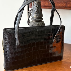 May include: A black, rectangular handbag with a crocodile skin texture. The bag has a zipper closure and two short handles. The handles are made of a smooth, dark material. The bag is sitting on a wooden surface.