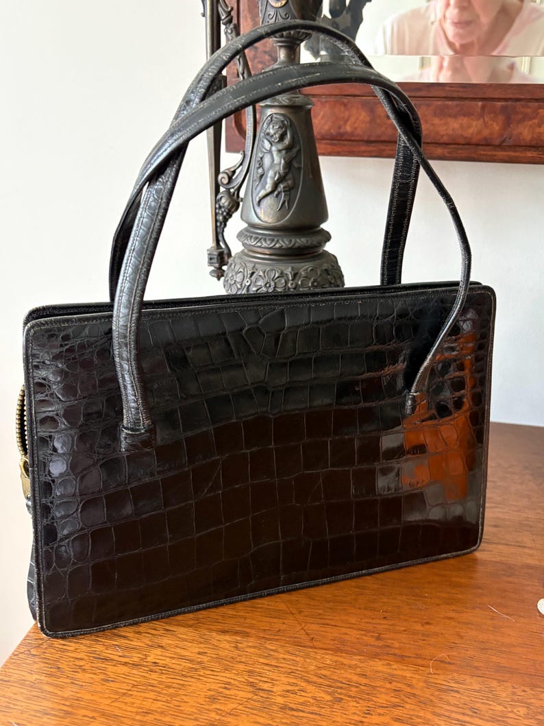 May include: A black, rectangular handbag with a crocodile skin texture. The bag has a zipper closure and two short handles. The handles are made of a smooth, dark material. The bag is sitting on a wooden surface.