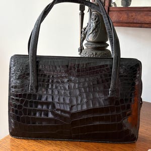 May include: Black rectangular handbag with a crocodile skin texture. The bag has two long, thin handles and a structured shape. The bag is sitting on a wooden surface.