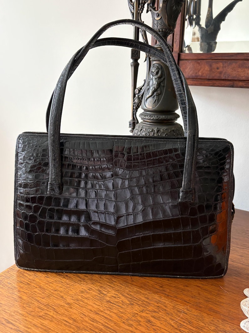 May include: Black rectangular handbag with a crocodile skin texture. The bag has two long, thin handles and a structured shape. The bag is sitting on a wooden surface.