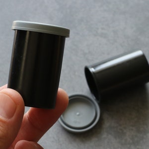 May include: Black film canisters with gray lids. One canister is upright, held by a hand, while another lies on its side. The lids are separate, showing the interior. These are used for storing 35mm film.