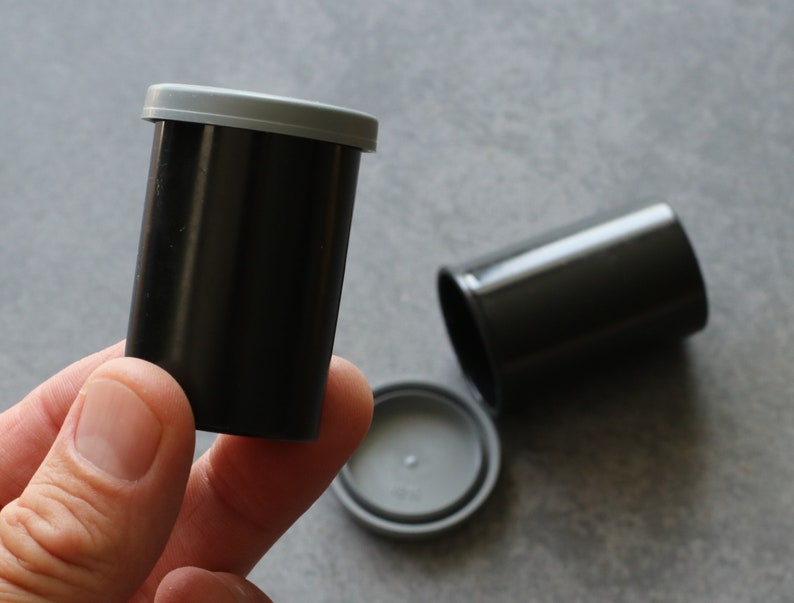 May include: Black film canisters with gray lids. One canister is upright, held by a hand, while another lies on its side. The lids are separate, showing the interior. These are used for storing 35mm film.