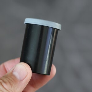 May include: A black film canister with a gray lid. The cylindrical container is held in a hand against a gray background. The canister is made of a smooth, glossy material.