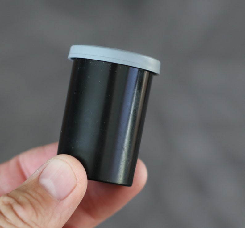 May include: A black film canister with a gray lid. The cylindrical container is held in a hand against a gray background. The canister is made of a smooth, glossy material.