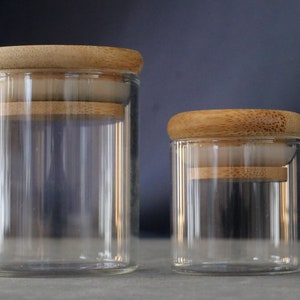 May include: Two clear glass jars with wooden lids. The larger jar is on the left and the smaller jar is on the right.