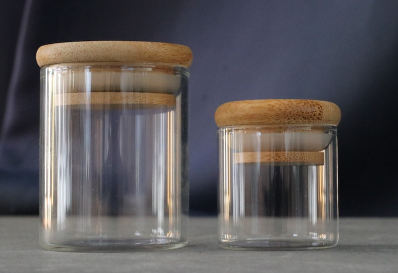 May include: Two clear glass jars with wooden lids. The larger jar is on the left and the smaller jar is on the right.