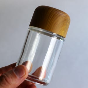 May include: A clear glass jar with a wooden lid. The jar is cylindrical and appears empty. The lid is a light brown color with a wood grain pattern. The jar is held by a person's hand against a plain background.