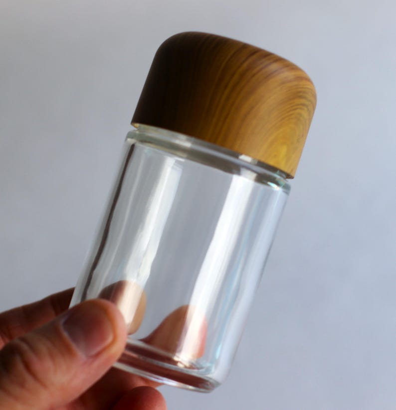 May include: A clear glass jar with a wooden lid. The jar is cylindrical and appears empty. The lid is a light brown color with a wood grain pattern. The jar is held by a person's hand against a plain background.