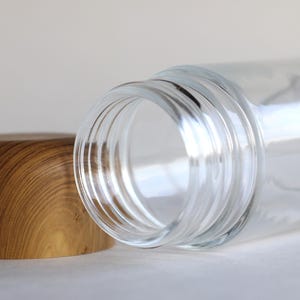 May include: A clear glass jar with a wooden lid. The jar is lying on its side, with the lid next to it. The lid is made of light brown wood. The jar has a wide opening and is empty.