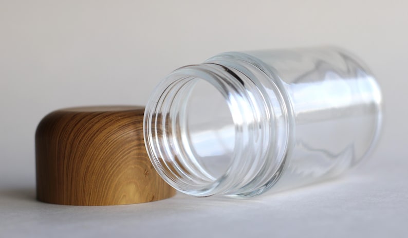 May include: A clear glass jar with a wooden lid. The jar is lying on its side, with the lid next to it. The lid is made of light brown wood. The jar has a wide opening and is empty.