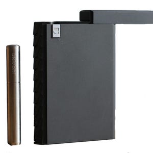May include: A black metal cigarette case with a textured exterior and a built-in tamper. The case has a hinged lid and a small, silver-colored tamper tool.