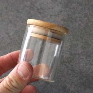 May include: A clear glass jar with a wooden lid. The lid has a light brown color and a smooth finish.