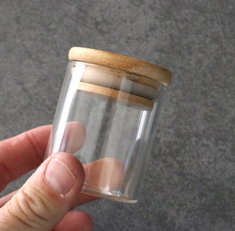 May include: A clear glass jar with a wooden lid. The lid has a light brown color and a smooth finish.