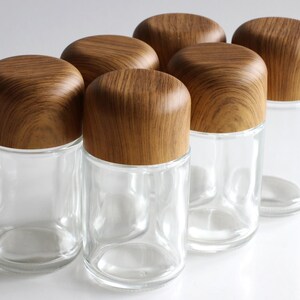 May include: Six clear glass jars with rounded wooden lids. The jars are cylindrical and have a simple, modern design. The wooden lids have a natural wood grain pattern. These jars are suitable for storing spices or other small items.