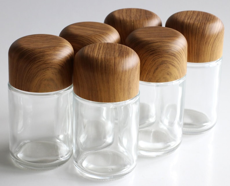 May include: Six clear glass jars with rounded wooden lids. The jars are cylindrical and have a simple, modern design. The wooden lids have a natural wood grain pattern. These jars are suitable for storing spices or other small items.