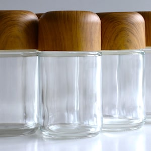 May include: Four clear glass jars with wooden lids. The jars are cylindrical with a rounded base and a wooden lid. The lids are a light brown color with a wood grain pattern. The jars are empty.