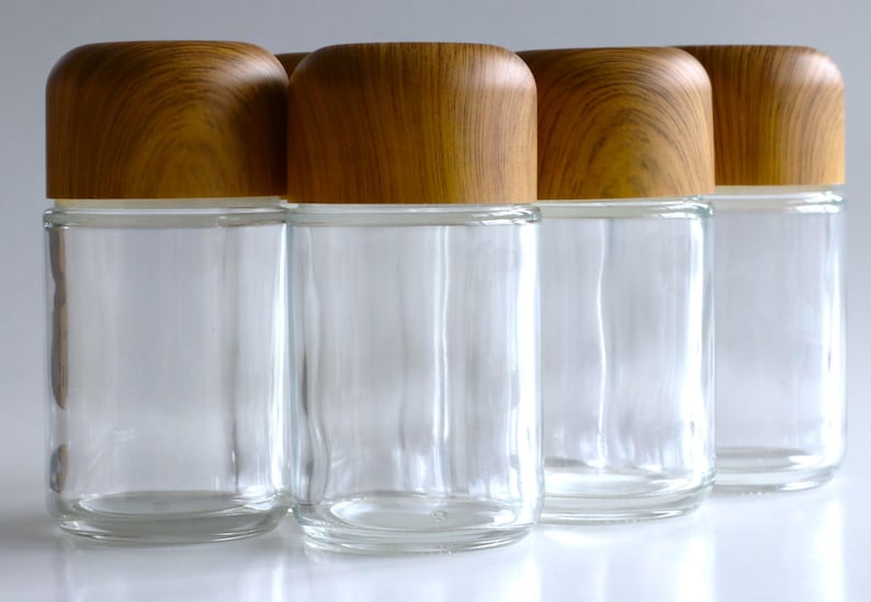 May include: Four clear glass jars with wooden lids. The jars are cylindrical with a rounded base and a wooden lid. The lids are a light brown color with a wood grain pattern. The jars are empty.