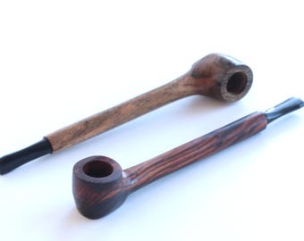 Op de afbeelding: Twee houten rookpijpen met lange stelen en zwarte mondstukken. De ene pijp heeft een lichtbruine, getextureerde houtnerf, terwijl de andere een donkerdere, roodbruine houtnerf met opvallende verticale strepen vertoont.