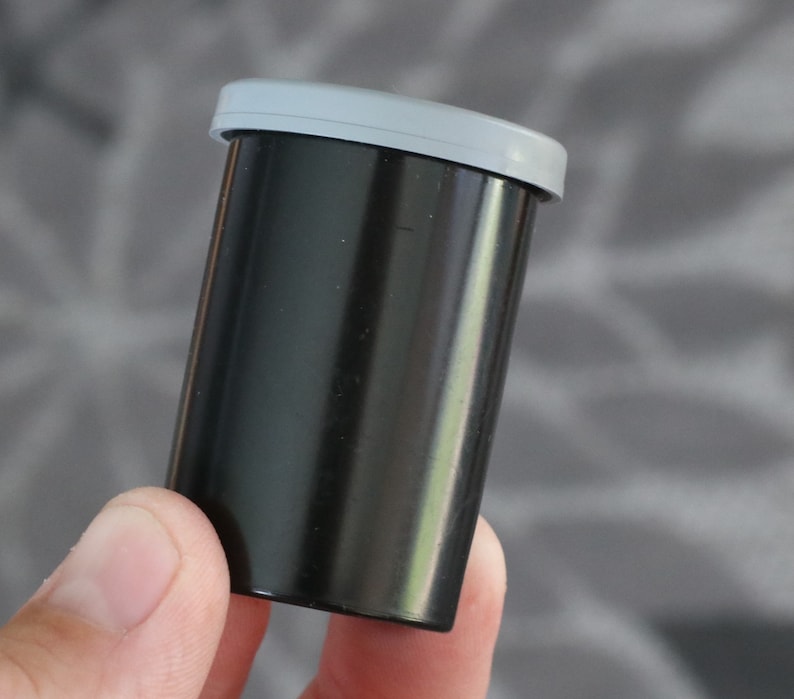 May include: A black film canister with a light gray lid. The cylindrical container is made of a glossy, dark material. The lid is a flat, oval shape.