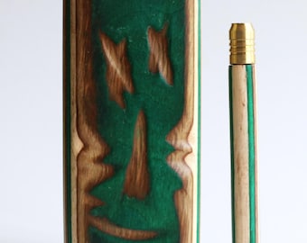 May include: A rectangular wooden case with a green resin inlay featuring a carved face design, accompanied by a matching cylindrical wooden pipe with a gold-colored metal mouthpiece. The wood grain is visible throughout the natural brown surfaces.