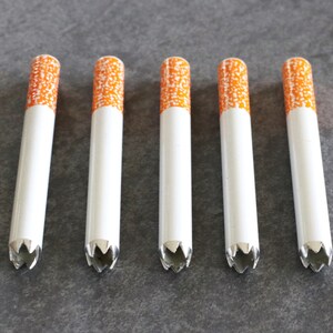 May include: Six white cigarette-shaped metal tubes with orange and white patterned tops. The tubes have a star-shaped opening at the bottom.
