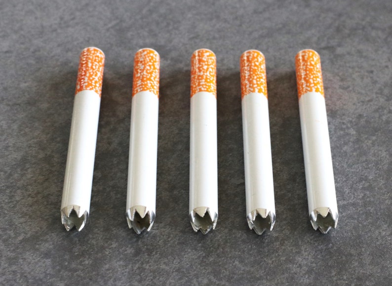 May include: Six white cigarette-shaped metal tubes with orange and white patterned tops. The tubes have a star-shaped opening at the bottom.