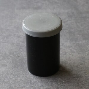 May include: A black film canister with a gray lid. The cylindrical container is made of a matte black material. The lid is a light gray plastic. The canister is sitting on a gray surface.