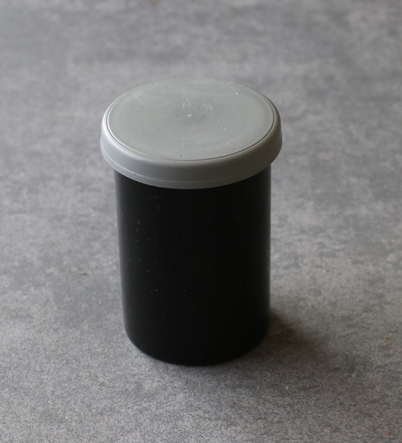 May include: A black film canister with a gray lid. The cylindrical container is made of a matte black material. The lid is a light gray plastic. The canister is sitting on a gray surface.