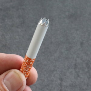 May include: A white cigarette tube with an orange and white patterned design. The end of the tube has a silver, star-shaped tip.