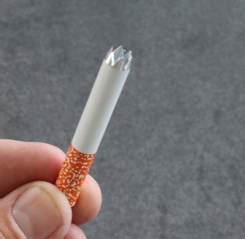 May include: A white cigarette tube with an orange and white patterned design. The end of the tube has a silver, star-shaped tip.