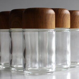 May include: A group of five clear glass jars with rounded, light brown wooden lids. The jars are cylindrical with a slightly wider base and are arranged closely together, reflecting light. The lids have a natural wood grain pattern.