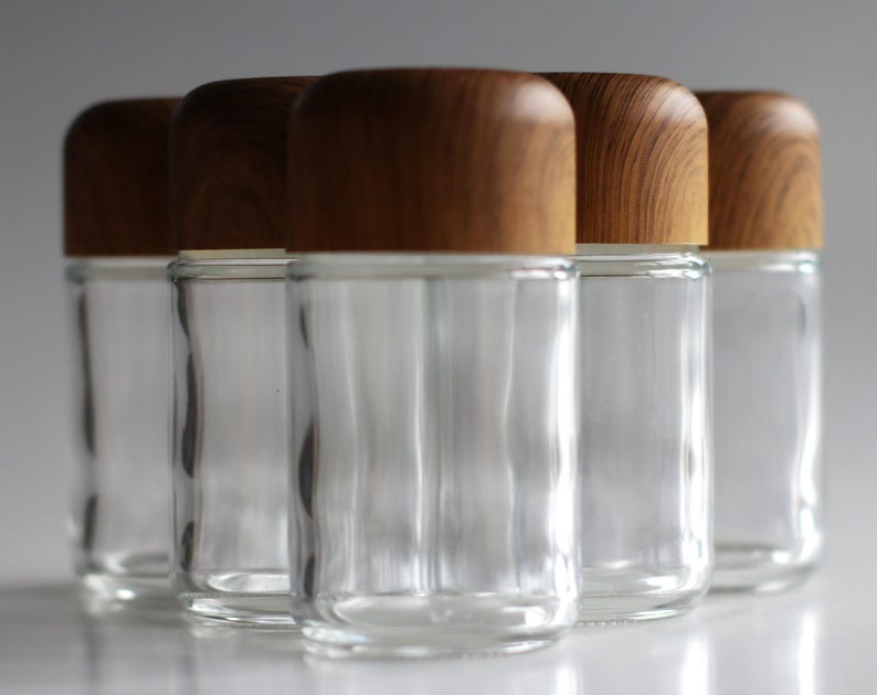 May include: A group of five clear glass jars with rounded, light brown wooden lids. The jars are cylindrical with a slightly wider base and are arranged closely together, reflecting light. The lids have a natural wood grain pattern.