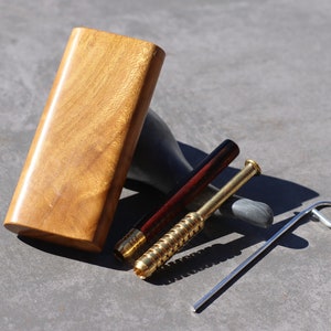 May include: A wooden dugout box with a golden finish, accompanied by a brass and wood pipe, a brass pipe, and a silver metal poker tool. The items are arranged on a gray surface.