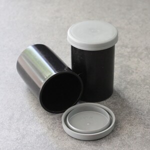 May include: Three black film canisters with gray lids. One canister is lying on its side, while another stands upright with its lid on. The third canister has its lid removed and placed next to it. The canisters are cylindrical.