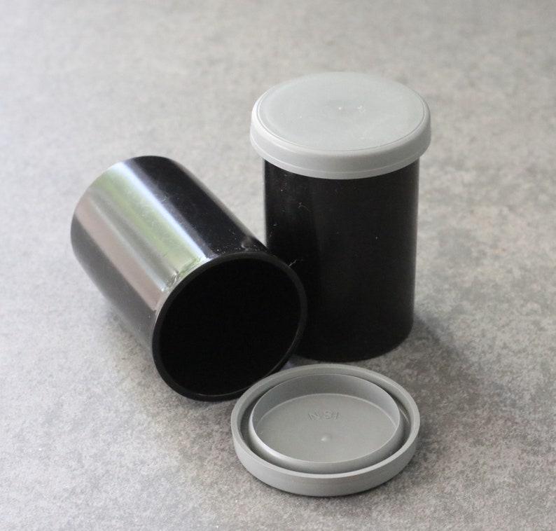 May include: Three black film canisters with gray lids. One canister is lying on its side, while another stands upright with its lid on. The third canister has its lid removed and placed next to it. The canisters are cylindrical.